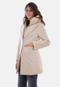 Cappotto beige a doppio strato con cappuccio rimovibile, caratterizzato da un materiale esterno strutturato, vestibilità sartoriale e tasche laterali. Accenti di design discreti.