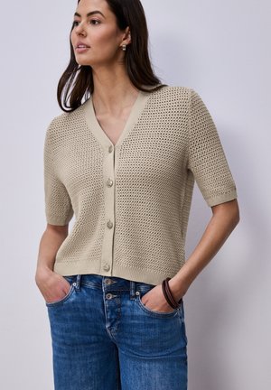 Femme portant un cardigan beige en tricot à manches courtes, boutonné avec un col en V, et un jean bleu, les mains dans les poches, regardant vers la gauche sur un fond uni.