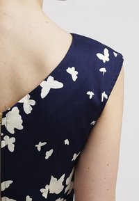 Marinblå klänning med v-ringning och kort ärm, prydd med vita fjärils mönster. Dragkedjedetalj längs ryggen. Slät textur.