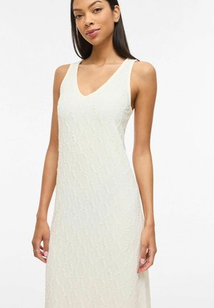 Mujer vestida con un vestido blanco sin mangas y de textura, con escote en V, posando sobre un fondo blanco liso.