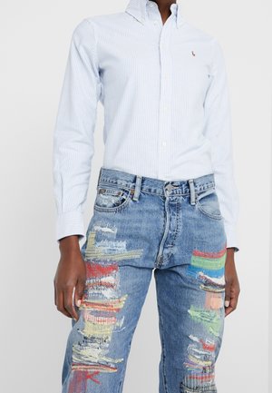 Osoba ubrana w jasnoniebieską i białą koszulę w paski, wpuszczoną w niebieskie dżinsy z kolorowymi łatami przypominającymi farbę i przetarciami.