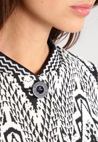 Svart och vit mönstrad tyg med en krage och en rund svart knapp som har varumärkesnamnet. Slät textur med synliga design detaljer.