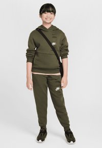 Dark green hoodie with adjustable hood and front pocket, paired with matching joggers. Both feature Nike logos in various sizes. Dark sneakers complete the look.