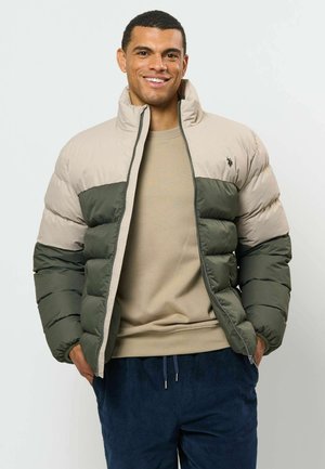 Homme souriant portant une doudoune beige et verte, un pull beige et un pantalon bleu foncé sur un fond clair uni.