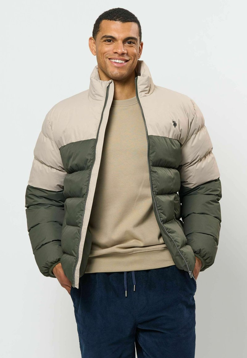 Homme souriant portant une doudoune beige et verte, un pull beige et un pantalon bleu foncé sur un fond clair uni.