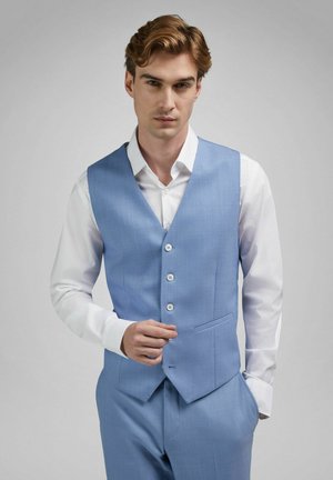 Uomo con capelli castano chiaro che indossa un gilet azzurro sopra una camicia bianca, con una mano in tasca e l'altra che sistema il gilet.