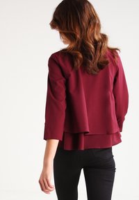 Blouse bourgogne à couches avec des manches trois-quarts, tissu doux et un ourlet arrière fluide, assortie à un pantalon noir ajusté.