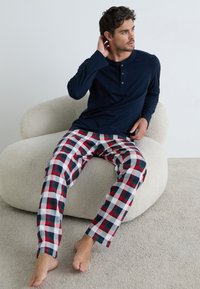 Man die op blote voeten op een beige moderne stoel zit, gekleed in een marineblauwe langwerpige henley shirt en een rood, groen, witte ruitbroek.