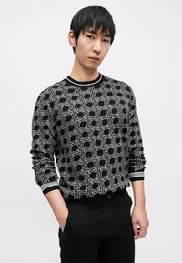 Juodas megztinis su balta geometrine rašto, apvalia iškilinta kaklo apdaila ir rankogaliais. Derinamas su juodomis kelnėmis.