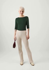 Green fitted top with three-quarter sleeves, paired with beige ribbed wide-leg pants. Holding a burgundy handbag and wearing cream-colored shoes.