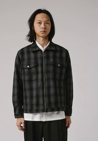Veste à carreaux noire et grise avec deux poches sur la poitrine, fermeture éclair à l'avant et col plat. Fabriquée en tissu doux et texturé.