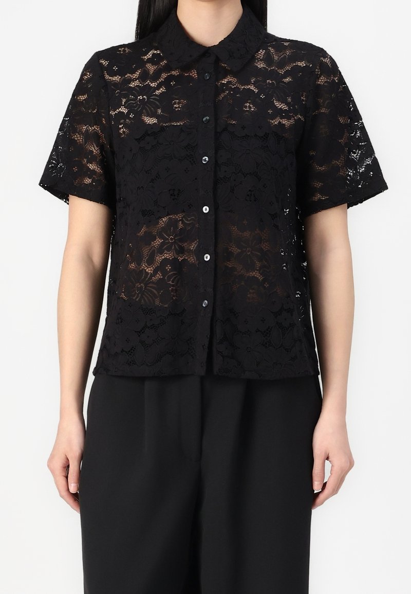 Femme portant un chemisier noir en dentelle à manches courtes, boutonné, avec des motifs floraux, et un pantalon noir large uni.