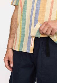 Korte mouwen shirt met horizontale meerkleurige strepen op een lichte achtergrond, met een merklabel aan de onderkant en een knoopsluiting.