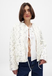 Veste matelassée blanche avec broderies florales vertes et violettes. Dotée d'une fermeture à cordon au niveau du cou et de poches latérales.