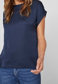 Světlomodrá saténová blůza s krátkým rukávem a kulatým výstřihem, hladká textura a uvolněný střih, v kombinaci se světlomodrými džíny.