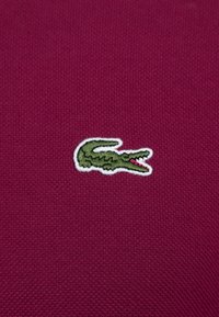 Tejido texturizado morado con un pequeño logo de cocodrilo verde contorneado en blanco, situado de manera prominente en la prenda.