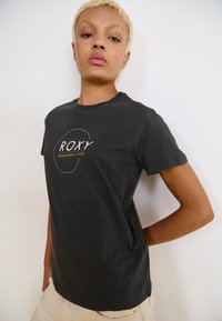 Juodas medvilninis trumpomis rankovėmis marškinėliai su apvalia iškirpte, su baltu ir geltonu "ROXY" logotipu ir minimalistiniu apvaliu dizaino akcentu.