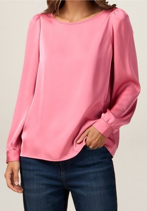 Femme portant un chemisier rose à manches longues avec des épaules légèrement bouffantes et un jean bleu, une main dans la poche, sur un fond uni.