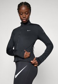 Femme portant une veste zippée noire Nike et des leggings ajuste la fermeture éclair latérale de sa veste, debout contre un fond gris uni.