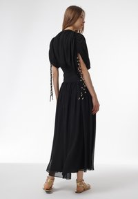 Schwarzes Kleid mit kurzen Puffärmeln, geraffter Taille und einem fließenden Rock. Verfügt über dekorative Bänder und Perlen an den Ärmeln. Getragen mit goldenen Sandalen.