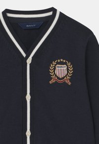 Tmavě modrý kardigan s V-výstřihem, bílým proužkovým lemem a emblémem GANT s vavřínovým věncem a detaily červené stuhy. Zapínání na knoflíky.