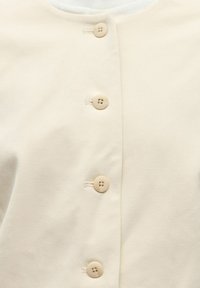 Gros plan de quatre boutons beiges attachés sur un tissu lisse de couleur crème clair, montrant les coutures et les boutonnières alignées verticalement.