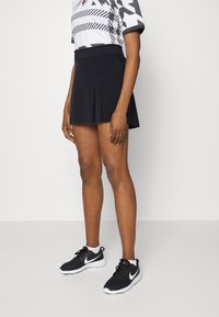 Černý plisovaný šortkový sukňový šortky vyrobené z lehkého materiálu, v kombinaci s vzorovaným šedobílým topem a černými teniskami Nike s bílými podrážkami.