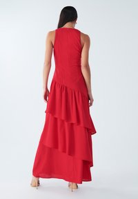 Czerwona sukienka maxi bez rękawów z wielowarstwowym falbaniastym wzorem, gładką fakturą, z dopasowanym gorsetem i zwiewną spódnicą. Zapinana z tyłu na zamek.