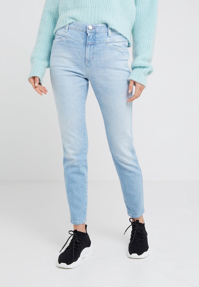 Personne portant un jean slim bleu clair, un pull doux bleu clair et des baskets noires à lacets avec des semelles blanches, debout sur un sol blanc.