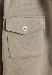 Prim-plan cu un material bej care prezintă o buzunar aplicat cu un buton cu clichet argintiu, textură netedă și cusături ordonate.