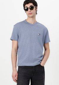 Μπλε βαμβακερό T-shirt με στρογγυλή λαιμόκοψη και κοντά μανίκια, διαθέτει ένα μικρό γραφικό λογότυπο στο αριστερό στήθος. Φοριέται με σκούρα τζιν.