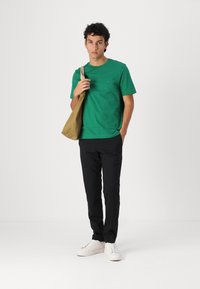 T-shirt en coton vert avec un col rond et un petit logo, associé à un pantalon noir et des baskets blanches. Tenant un sac en toile beige.