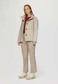 Beżowy płaszcz wełniany z wysokim kołnierzem i pięcioma guzikami, zestawiony z spodniami w kratkę w kolorze kremowym, brązowym i czerwonym. Buty w jasnym beżu z masywną podeszwą.