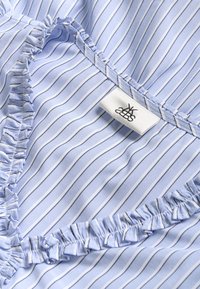 Material bleu deschis cu dungi albe înguste, având o margine cu volane și o etichetă cu text negru. Textură netedă cu un luciu ușor.