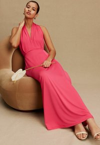 Lange roze halterjurk met een getailleerd lijfje, vloeiende stof en een lichte glans, gecombineerd met witte sandalen met bandjes. Zittend op een ronde beige stoel.