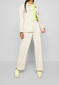 Beige denimjacka med guldknappar, matchad med en neongrön skjorta och beige vida byxor. Klädkombinationen fulländas av klara gula sneakers.