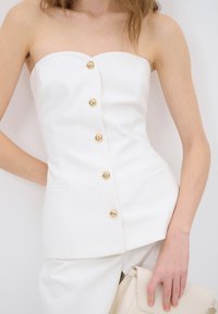 Femme portant un haut blanc sans bretelles avec cinq boutons dorés, un pantalon blanc assorti, tenant un sac à main beige sur un fond clair.