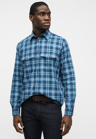 Άνδρας με μπλε και πράσινο καρό πουκάμισο με τσέπες στο στήθος και σκούρο τζιν, κοιτάζοντας προς τα αριστερά του με φόντο απλό.