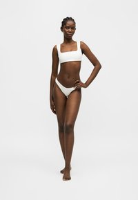 Γυναίκα μοντέλο ποζάρει με λευκό δίκομψο μαγιό με τετράγωνο λαιμόκοψη και ψηλόμεσο κάτω μέρος σε απλό ανοιχτό φόντο.