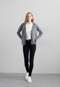 Grauer Cardigan mit Knöpfen, getragen über einem weißen Hemd, kombiniert mit schwarzen Slim-Fit-Hosen und weißen Sneakern. Glatte Textur, lässiges Design.