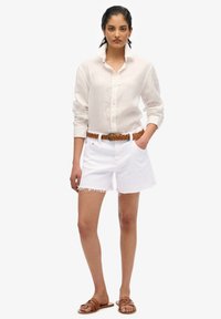 Chemise blanche en lin à boutons, short en denim blanc effiloché et ceinture tressée beige. Le modèle porte des sandales marron. Tenue d'été simple et décontractée.