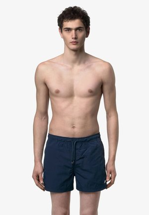 Giovane uomo con capelli corti e scuri che indossa pantaloncini da bagno blu navy, in piedi di fronte a uno sfondo chiaro e uniforme.