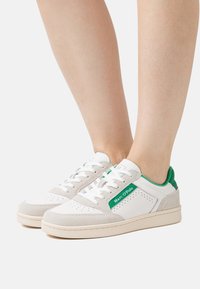 Białe i beżowe sneakersy z zielonymi akcentami oraz logo "Marc O’Polo" noszone na bosych stopach na jednolitym tle.
