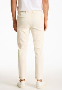 Pantaloni di colore crema chiaro con una vestibilità slim, dotati di tasche posteriori e risvolti, abbinati a sneakers bianche su uno sfondo neutro.