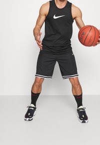 Homme portant un débardeur Nike noir, un short et des baskets Nike, tenant un ballon de basket orange, debout devant un fond uni.