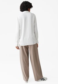 Weiße Bluse mit langen Ärmeln und knöpfbarem Manschetten, kombiniert mit lockeren beigen Hosen und weißen Sneakern mit schwarzen Akzenten. Glatte Textur des Stoffes.