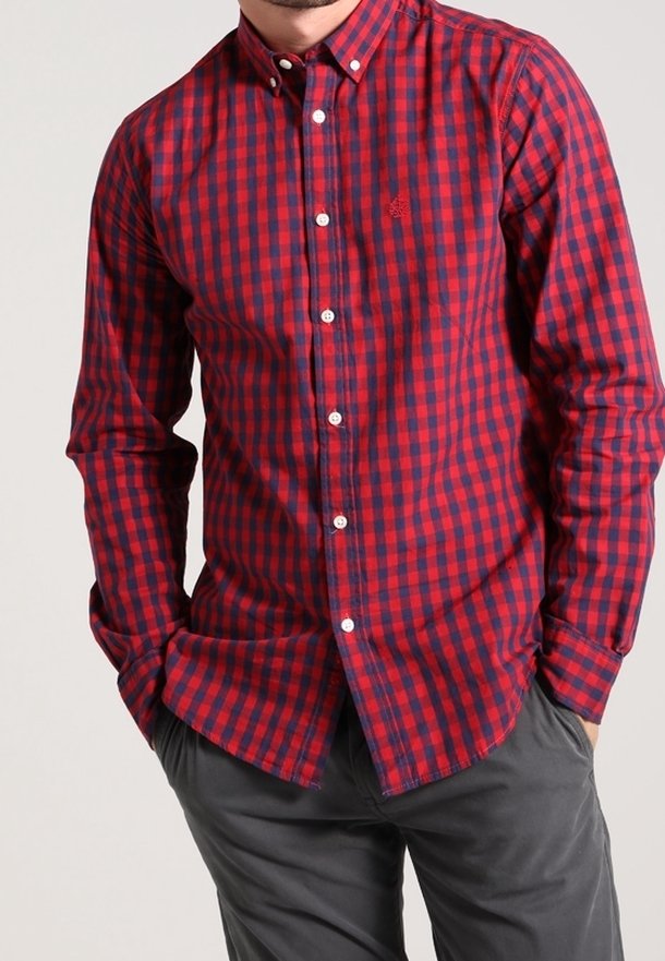 Homme portant une chemise à carreaux rouge et bleu marine à manches longues et un pantalon gris, mains dans les poches, sur un fond clair uni.
