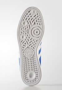 Vit sula på sneakers med blå accenter. Har ett texturerat greppmönster med cirkulära och geometriska former. Varumärkets logotyp vid hälen.