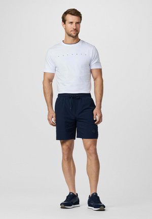 Man draagt een wit T-shirt met korte mouwen en het logo "Hackett", marineblauwe short met trekkoord en donkerblauwe sneakers, staand tegen een effen achtergrond.