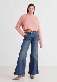 Pink cropped sweater with ribbed turtleneck, paired with high-waisted, flared blue denim jeans. Model wearing beige heels.